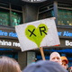 A protester raises an Extinction Rebellion sign as a ticker in the background displays a story about blackouts in California in the face of wildfire risks.